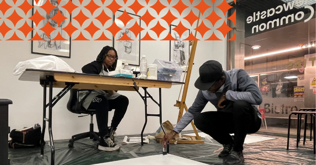 Artists in residence at Newcastle Common as part of the TRIBES: Matabeleland to Staffordshire exhibition. Two people work in an open space. One is sitting behind a s desk sketching. The other is crouching down in front of the desk preparing a canvas with a base layer of paint.