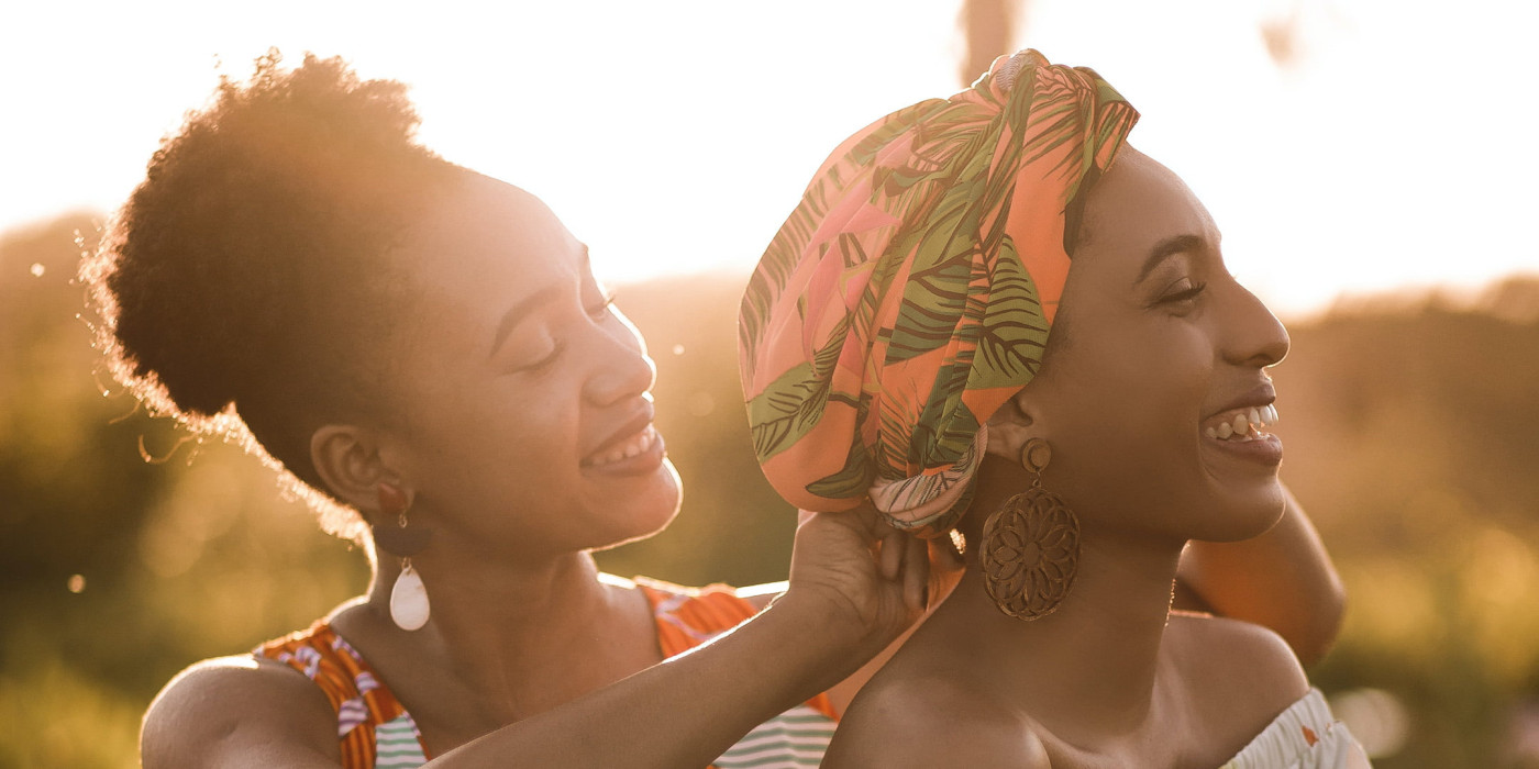 Two women are pictured from the shoulders up. One stands behind the other and helps to wrap a brightly coloured head covering. They are both smiling and clearly happy.