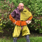 A jeser in coulourful medieval style clothes poses with hands on hips. His head is lowered pulling a funny face. He is bald and has a dark moustache. He stands in front of a hedge on some grass.