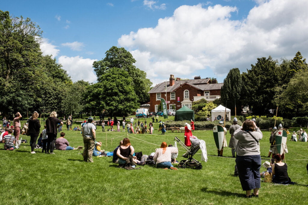 Brampton museum is shown in the distance with a large grassed area in the foreground. People are on the grass and there are some gazebos. Two knights have their back turned revealing brightly coloured shields.
