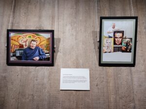 Twp potographs hang against a light-coloured wood effect wall. The image on the left is in landscape format and shows a man in a blue jumper sitting at a desk in front of a fairground sign. The image on the right depicts a man standing behind some large pieces of fairground art of Robbie Williams, Britney Spears and Lady Gaga.