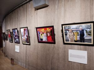 Coloured photographs are displayed against a light wood effect wall. The wall curves around to the left so that the line of photographs disappears from view. Spotlights shine down of the photographs from above.