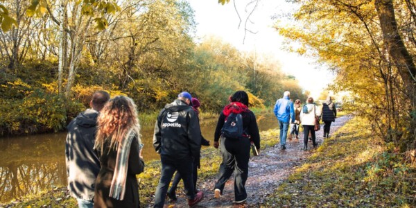 The group of walkers are seen from behind walking along the canal towpath. The canal is to their left and trees, foliage and grass surround the scene to both sides.