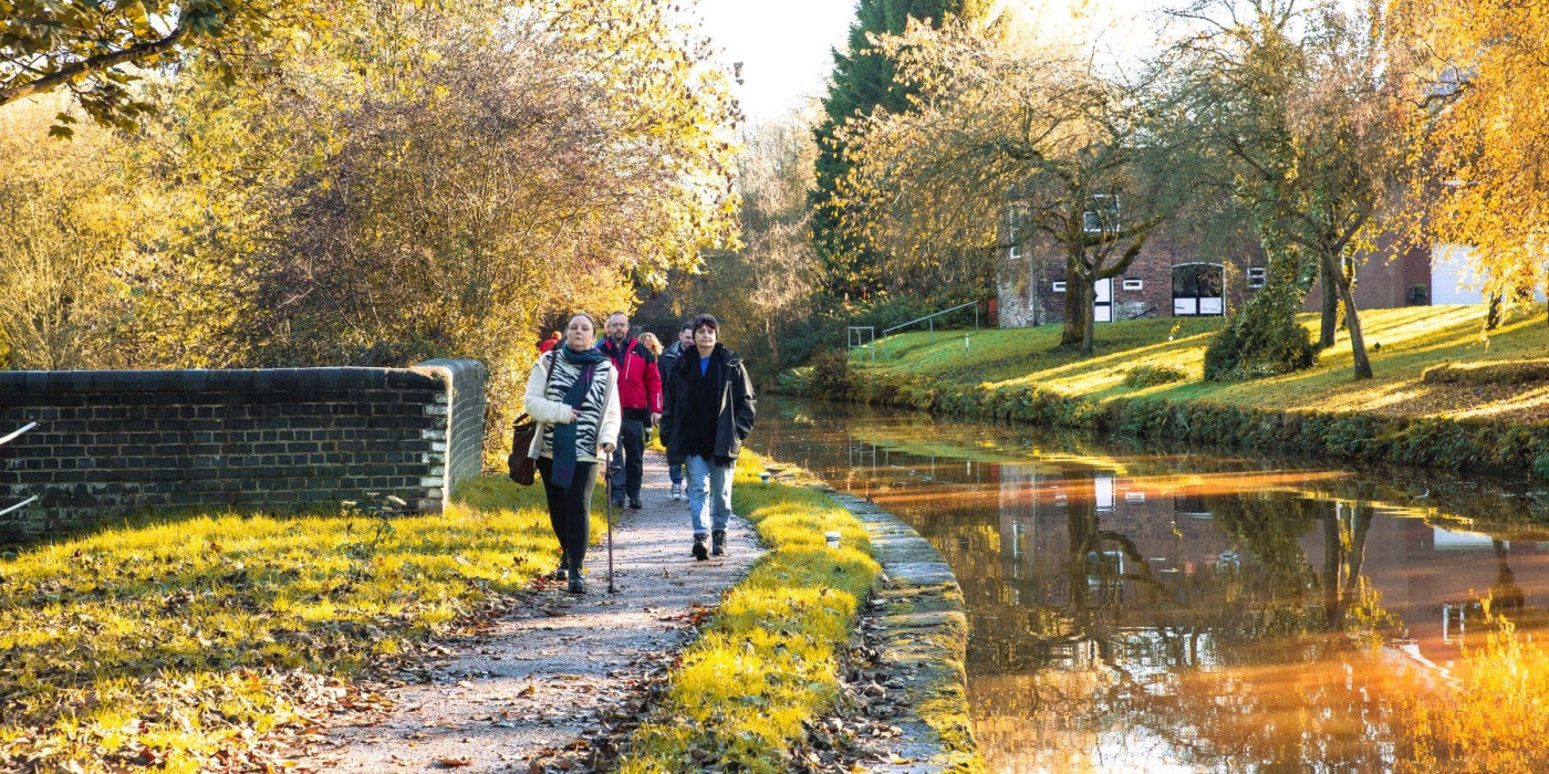 The group of walkers are coming towards us in a line along the towpath. To the right is the canal and on the opposite bank are trees, a grass slop and a building in the background. The trees and building are reflected in the sunshine onto the orange water.