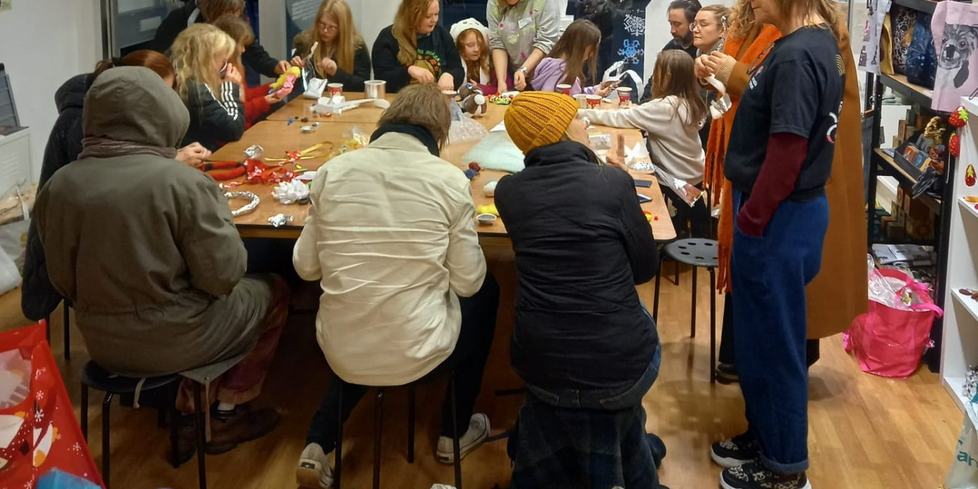 A group of people sit around a large square table. Behind them is a window with the name Newcastle Common seen against a dark sky. In the foreground are brightly coloured craft materials on a table.