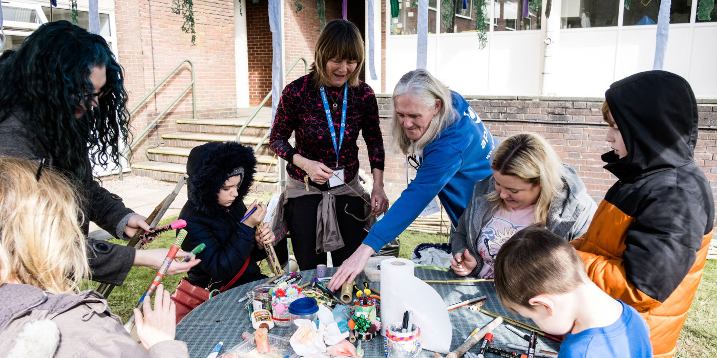 A group of adults and children are gathered around a table outdoors. On the table in front of them there are paints. There is a person wearing a blue Appetite lanyard and a Appetite volunteer wearing the blue Appetite uniform is leaning across and helping the people taking part in the activity.
