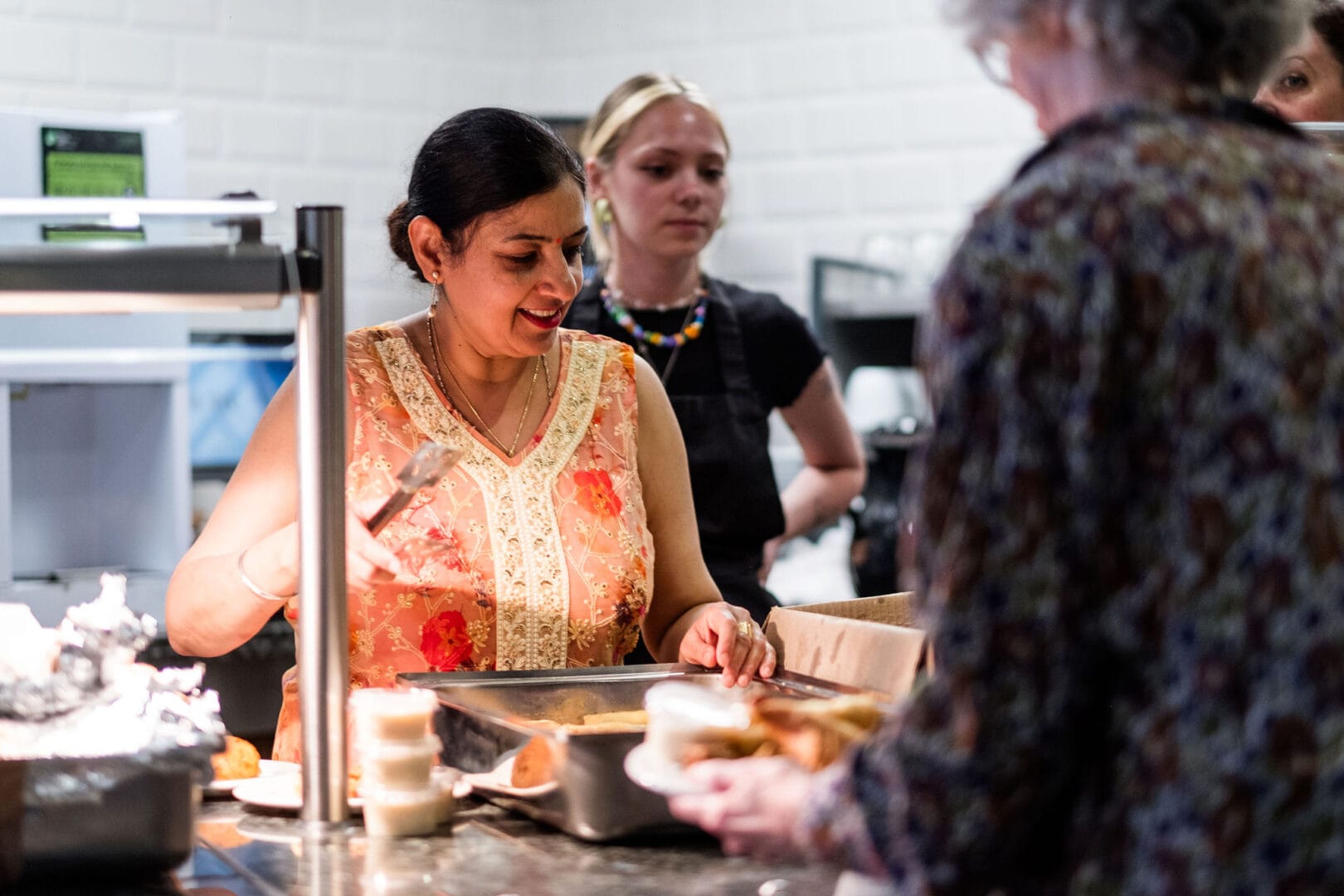 A lady in an orange and yellow sari-style dress is standing behind the food counter at the New Vic Café. She is serving somebody whose back is to us with food from a large silver tray.