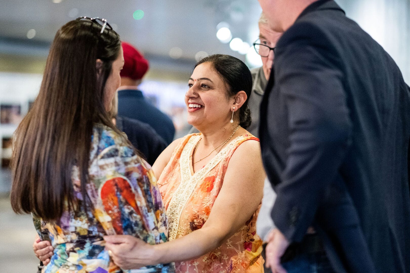 Two women are in conversation. The lady on the right is wearing a yellow and orange sari dress with a red bindi. She is facing her friend, holding her by the arms and smiling broadly. The lady on the left can be seen mostly from behind and is wearing a multicoloured abstract shirt and has glasses perched on the top of her head. Two men stand around them but we can’t see their faces.