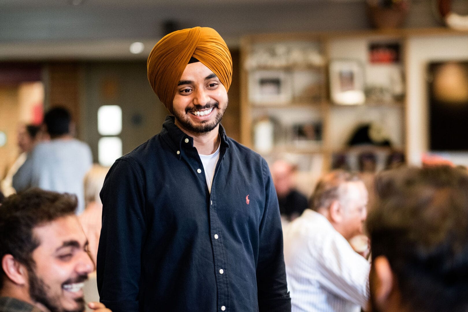 A man in a dark blue shirt with a red Ralp Lauren logo and a mustard yellow turban is standing in front of a group of seated people. He has a big smile on his face. To the left we can see one of the seated group in profile laughing. Other crowd members can be seen in the background of the New Vic Theatre café area.