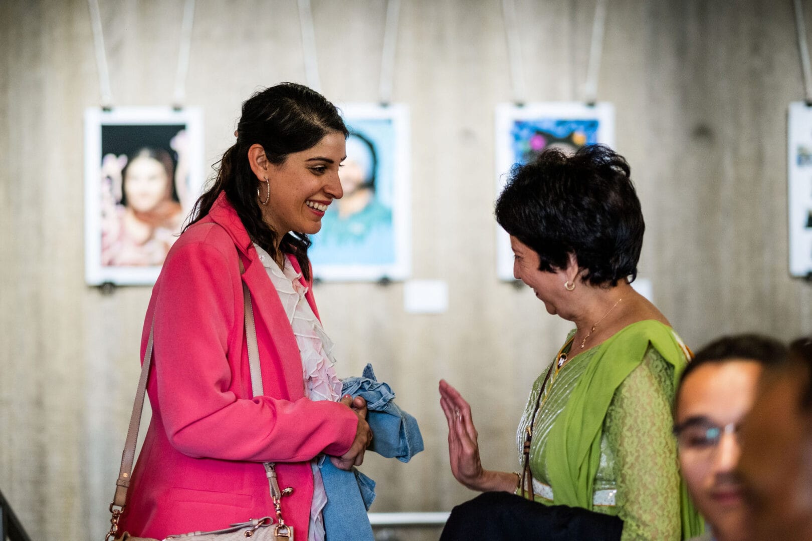 Two women are standing and talking. On the left, one woman wears a bright pink blazer with a cream ruffled top, and a blush shoulder bag. The lady on the right is in a bright green sari and is holding the palm of her right hand up as she speaks. Both ladies are laughing. In the background you can see blurred images of photographic portraits hanging in front of the wall.