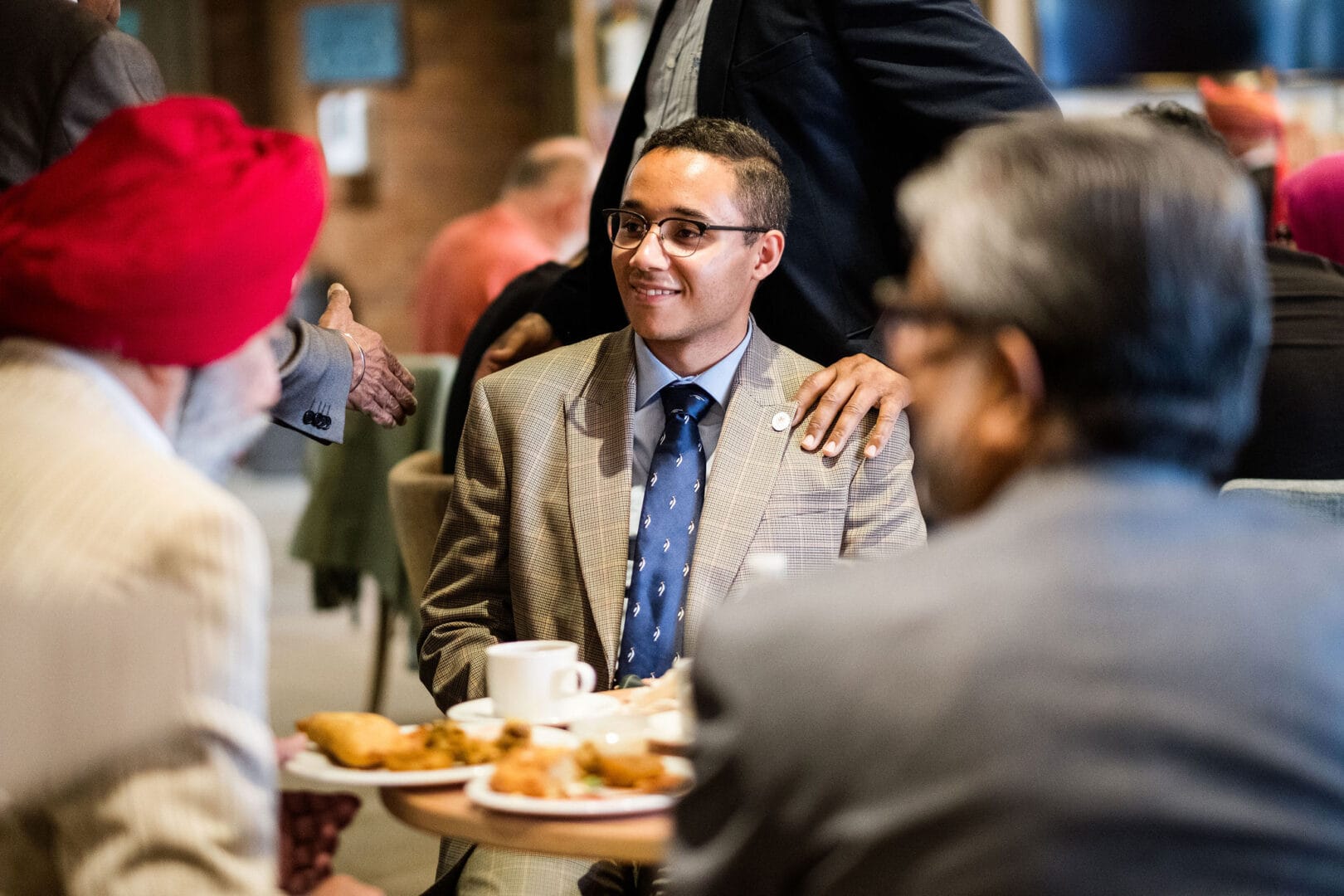 A closeup of a group or people, three seated and two standing. We see the back of two men sitting down who are both wearing suits, one with a red turban. They are facing a young man also in suit and tie who is smiling. Behind him, two men are standing, but we can’t see them above shoulder height. One man has his hand resting on the shoulder of the young seated man, while the other holds out his hand for a shake.