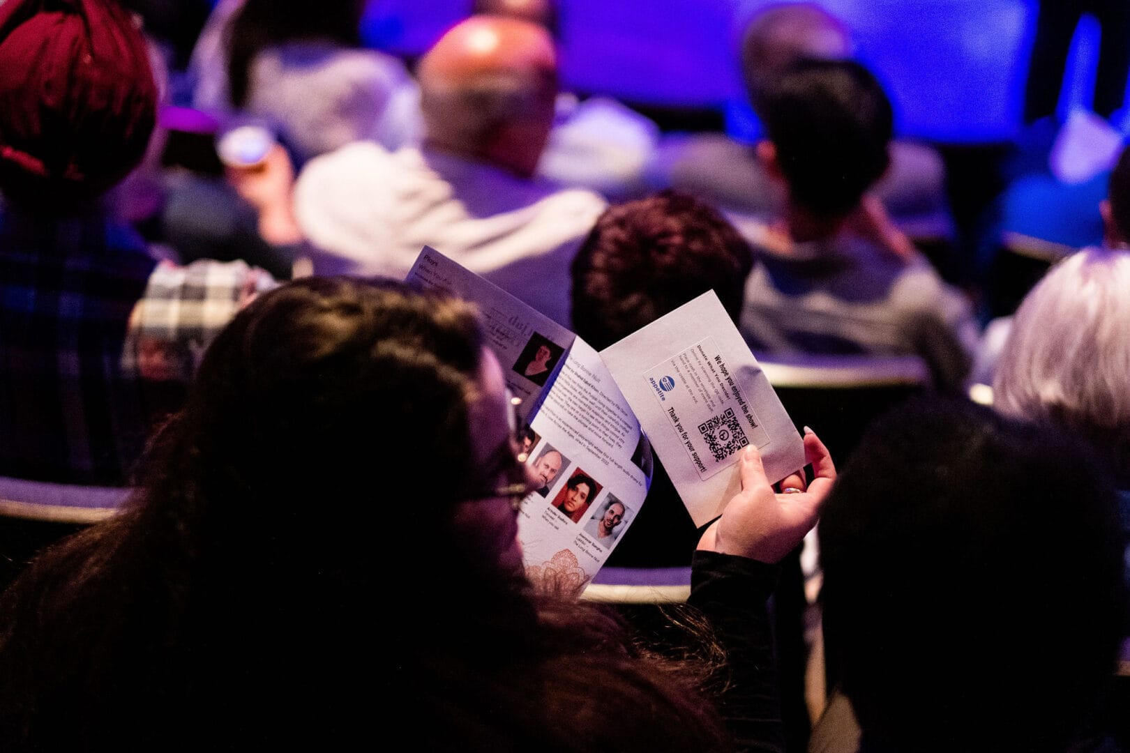 We are looking over the shoulder of a woman in an audience. She has long black hair and glasses that can be seen as she’s facing to her right. In her left hand she is holding a booklet that shows pictures of actors and one of the playwrights along with some text. In her hand she holds an Appetite donation envelope that says: “We hope you enjoyed the show! Donate what you decide. Thank you for your support.”
