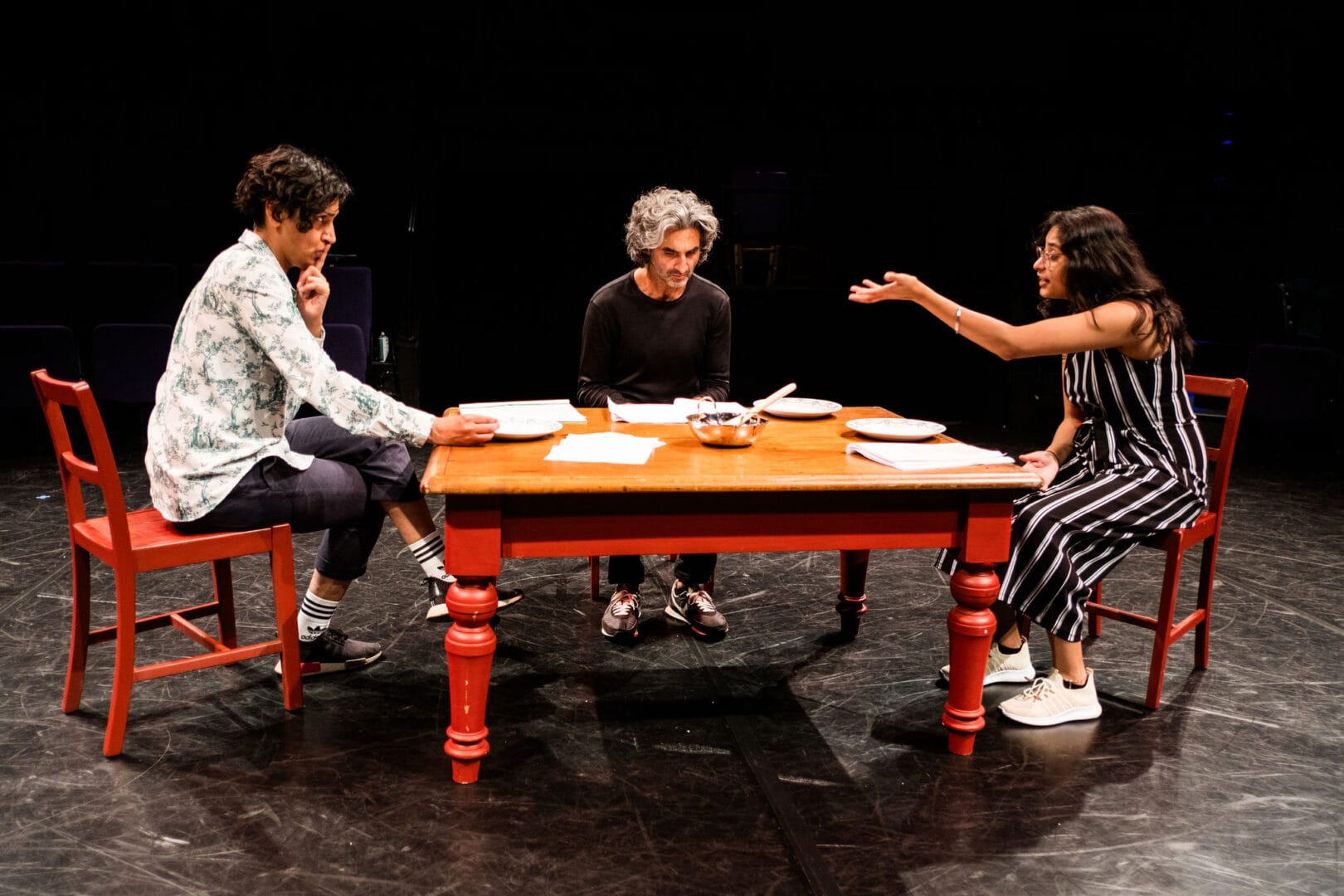 A man, woman and young woman are sitting on stage around a table with red legs. The background is completely black. On the left-hand side, the woman sit on a red chair. She is casually dressed in jeans, shirt and trainers. She is making a shushing gesture towards to young woman opposite with a finger to her lips. In the middle, the man, dressed all in black, is looking down at some papers on the table. On the right, the young woman is gesturing to the woman opposite with her left hand in the air. She has a black-and-white jumpsuit and glasses. On the table are plates, a bowl and mixing spoon, and papers.