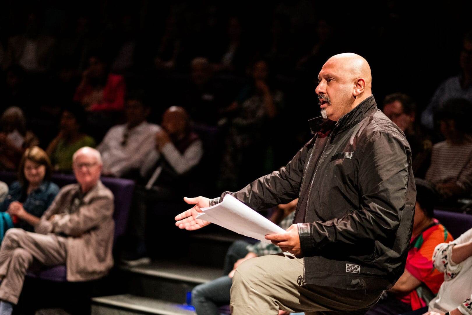 A man is standing in front of a seated audience. He can be seen from hip level upwards, but you can tell that he has one foot propped up on something and his knee is raised. He is gesturing out towards the side with his right hand. He’s holding a script looking at and speaking to somebody off screen to the left.