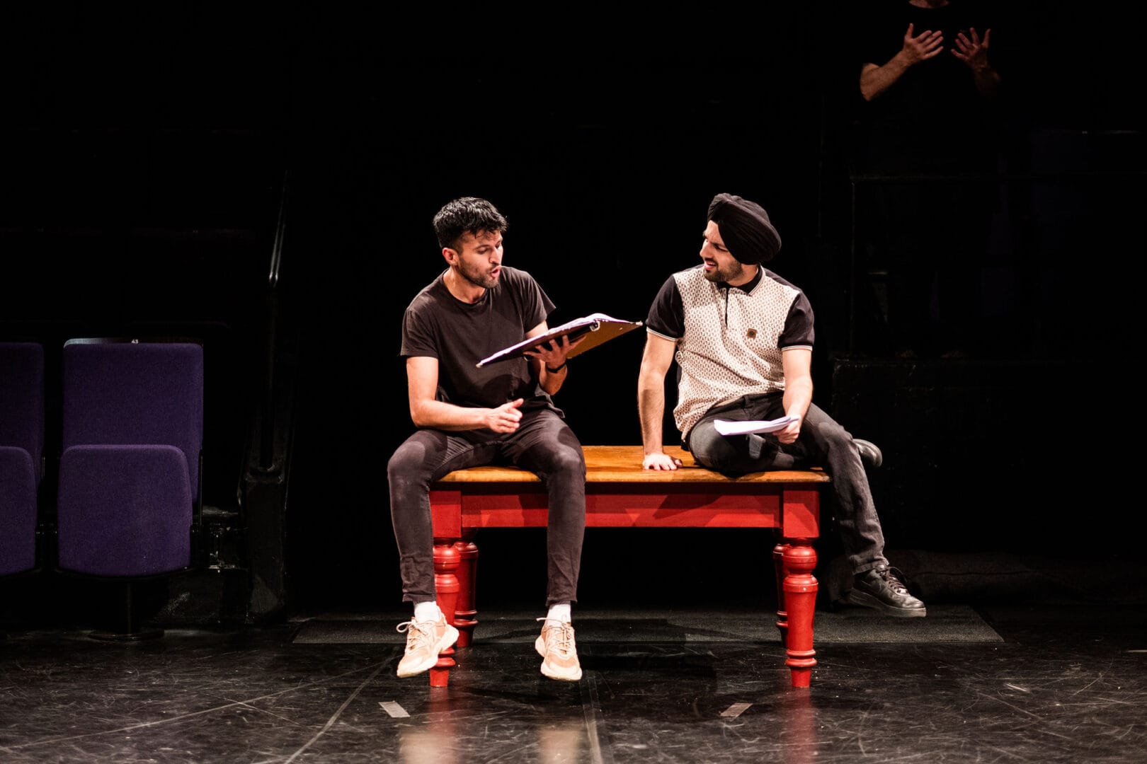 Two young men casually dressed in jeans and trainers are sitting on stage on a table with red legs. The backdrop is solid black. The man on the right wears a black turban, while the other wears none. One is speaking, consulting a script that he holds while the other looks at him.
