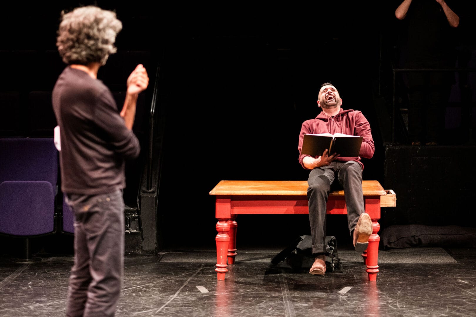 Two men are on a stage. The background is solid black. One man is seen from behind looking toward the other, who is sitting on a table with red legs. The seated man is holding a script and throwing his head back in laughter.
