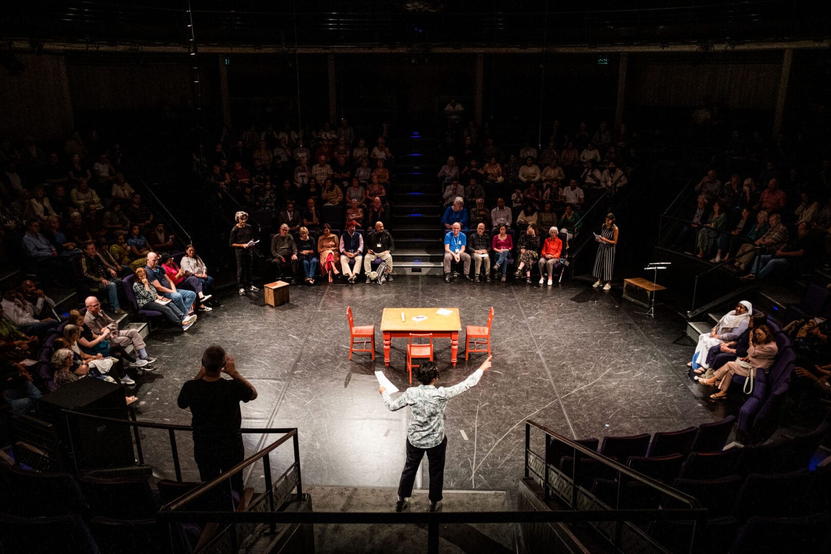 A theatre auditorium in the round is shown from an elevated perspective from the top pf a set of stairs. The seating audience are in the dark but you can see people on the front row quite clearly as they encircle the central stage. In the centre of the stage is a wooden table with red legs and three red wooden chairs arranged around it. We can see an actress from behind speaking to the audience. In her left hand she holds a script and her right is raised and pointing. To her left we can see a man dressed in black providing sign language.