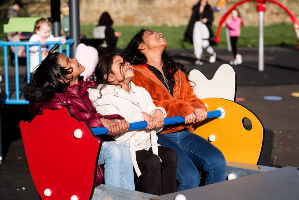 Three children are sat on a seat on a park. They are smiling, their heads turned up to the sky to face the sun.