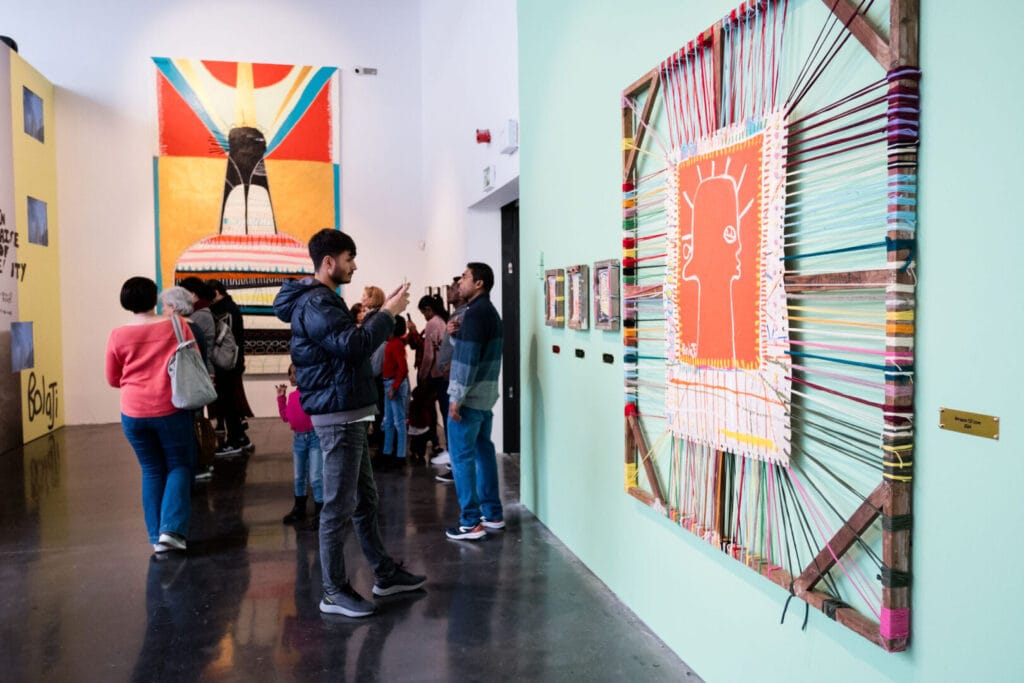 A group of people are looking at artwork in a gallery. One person is stood taking pictures of the artwork on a smartphone,