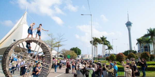 Five performers are alongside a large, wooden wheel. Two are stood on top of the wheel, one is climbing inside and two are stood either on the ground. They are outside and there is an audience all around, watching and holding up phones and cameras.