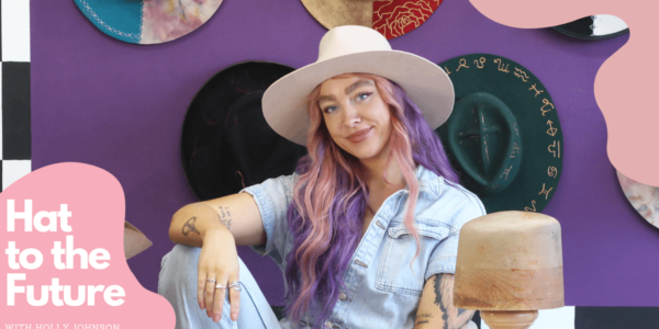 A person with long pink and purple hair is looking into the camera. They are wearing a white hat and a denim boiler suit. They are resting their right arm on their right knee and are sat behind a desk. On the desk is a hat block and other tools. Behind them is a purple wall, with lots of different hats hung up. There are spink splodges in the top right and bottom left hand corners. The bottom right has white text which reads 'Hat to the Future with Holly Johnson'.