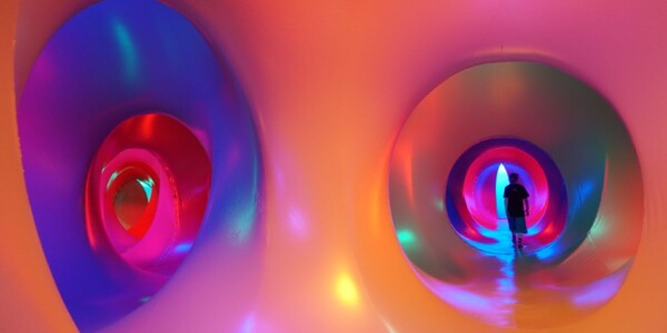 We are looking at the interior of a Luminarium, an inflatable structure with corridors and chambers bathed in coloured light. From this view we are standing in the intersection of two corridors that can both be seen down. The corridors are circular and oval in shape and undulate side to side as they progress. The foreground where we are is a soft peach colour, and you can see the colours morph and blend as your eye travels down the corridors into blues, pinks and reds with purple and green spotlights at points adding pattern. You can see the black silhouette of a lone figure walking down the end of corridor on the right.