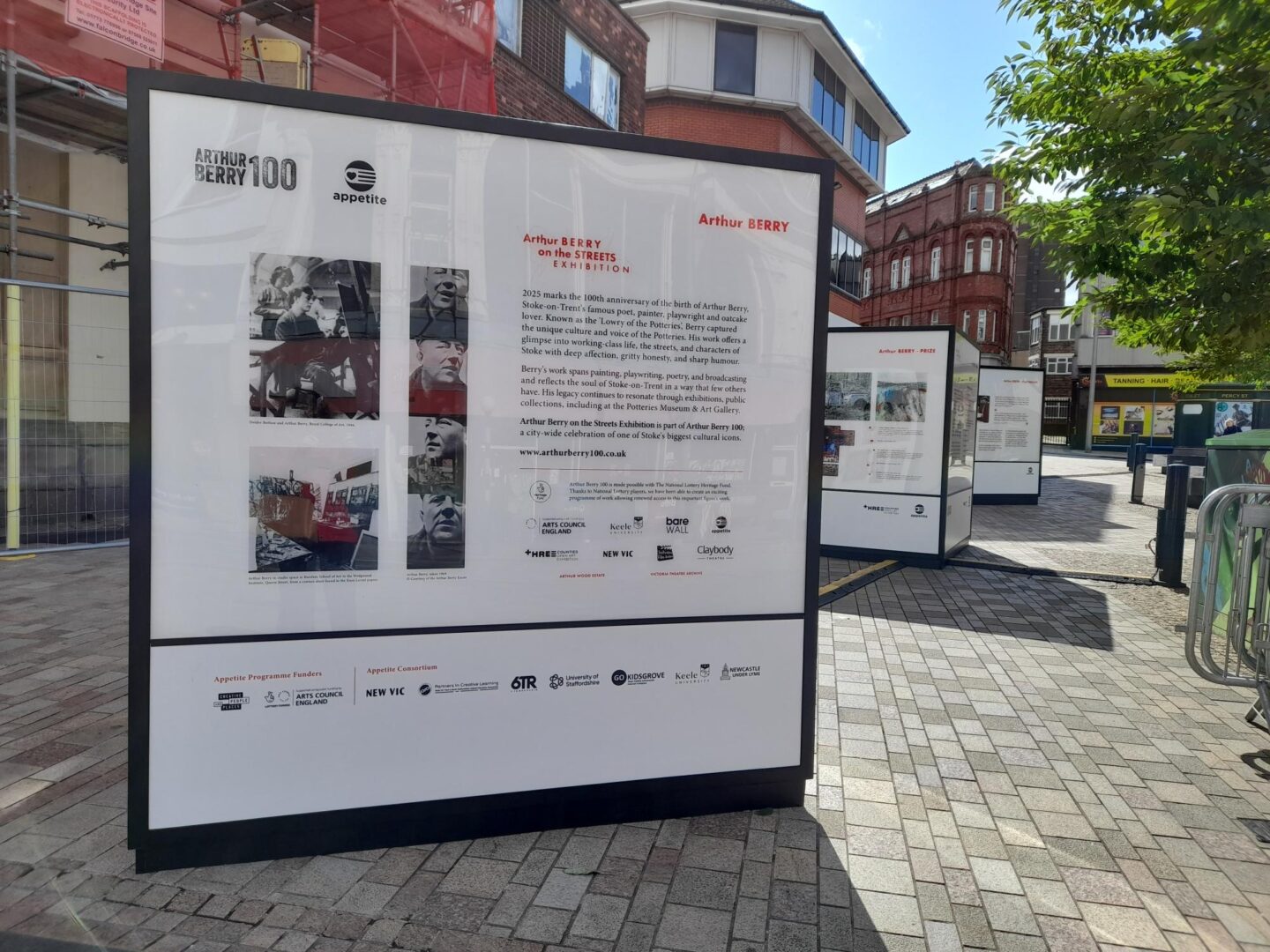 The image shows a street on a sunny day. The focus of the picture are three large cubes that contain writing and images on about Arthur Berry, creating an outdoor exhibition.