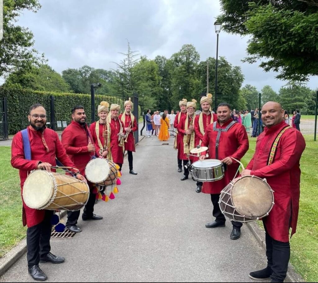 Bombay Baja: Two rows of men in long red tunics are lining a path in a park and looking towards camera. The ten men vary in age and ethnicity. The four men closest to camera have drums while the six towards the rear have brass instruments and golden turbans.