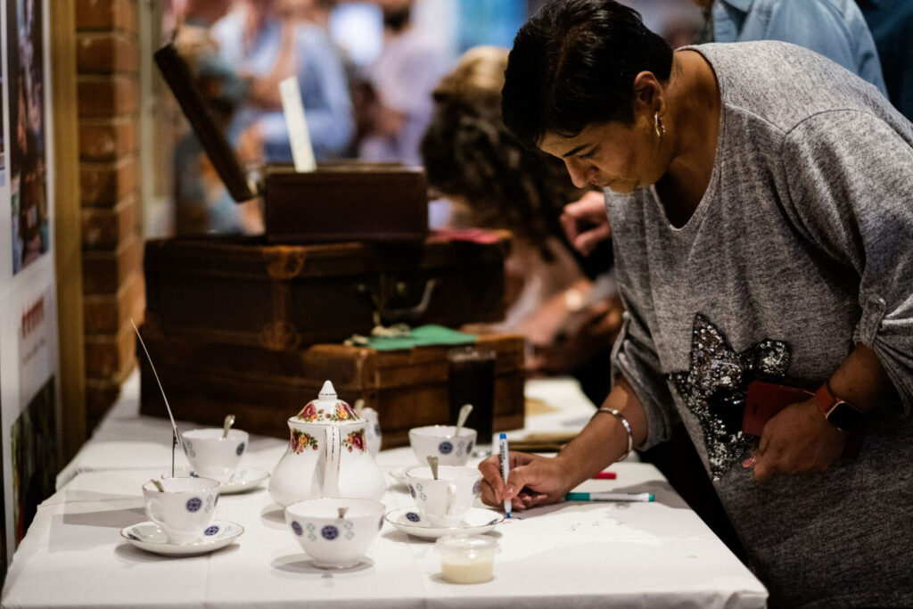 A South Asaian with short, black hair and wearing and loose-fitting, grey top with a sparkly butterfly on is leaning over a table. On the table are a china tea set and set of three brown suitcases stacked. The lady is holding a market pen and writing on some paper on the table. Blurred images in the background suggest that she is in a busy area.