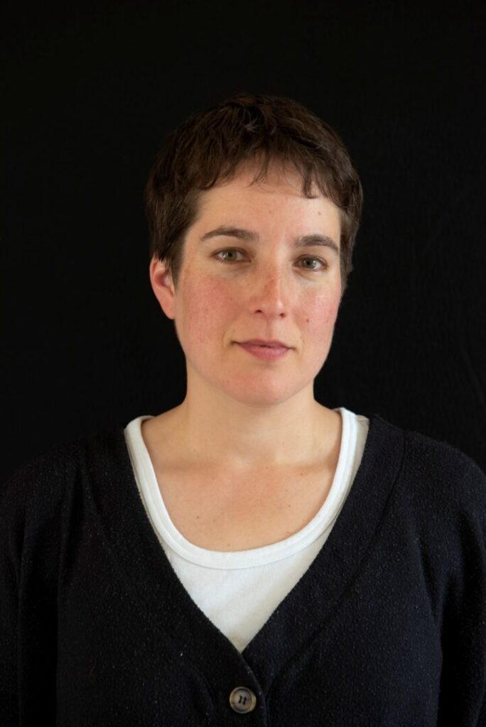 A white person is facing the camera against a black background. They have short brown hair and a slight smile in their eyes. The portrait is head and shoulders only nd as they are wearing a white v-neck t-shirt and black v-neck cardigan with brown buttons, the effect is striking against the dark background.