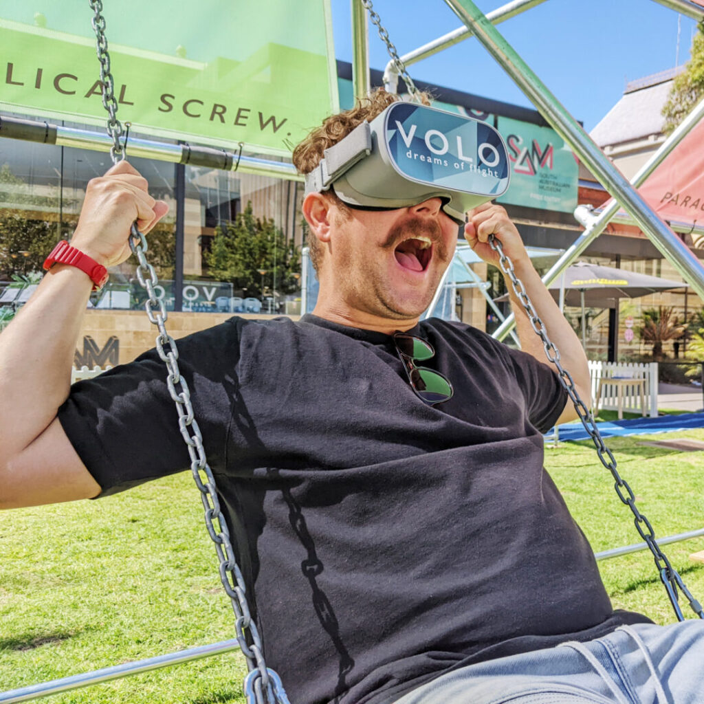 A man is seen from the waist up swinging on a swing with metal chains. He’s surrounded by a structure that we can partly see that consists of metal poles and pastel-coloured Perspex panels with writing on them. The man has a moustache and is wearing a black t-shirt with sunglasses tucked into the neck. He’s also wearing a large pair of grey goggles that have a blue panel over the eye area with words on it reading ‘VOLO dreams of flight’. The man has his mouth wide open in a way that looks like he’s enjoying what he can see in the goggles.