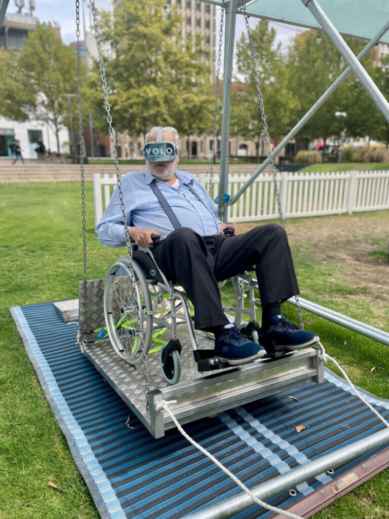 An older man with white hair and beard is in the open air on a patch of grass. He’s sitting in a wheelchair that is on a metal platform attached to swing chains and surrounded by a structure of metal scaffolding that we can’t see in full. The man is wearing a large pair of grey goggles that have a blue panel over the eye area with words on it reading ‘VOLO dreams of flight’.