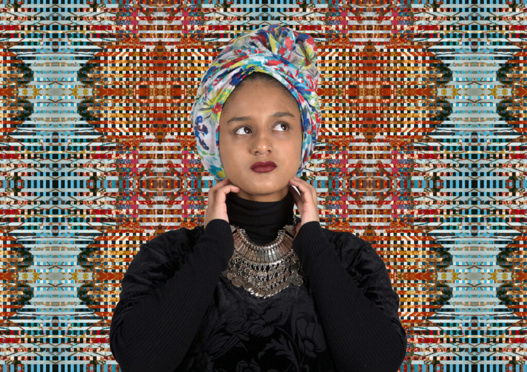 Nilupa Yasmin - a young woman of South Asian heritage - is seen head and shoulders standing in front of a colour artwork that fills the background. The art is of many thin strips of pattern and colour in primarily red and blue colours interwoven. She wears black clothing with a turtle neck sweater, a colourful head wrap and a deep red lipstick. A large silver necklace stand out over her dark clothing.