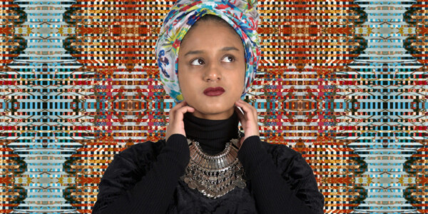 Nilupa Yasmin - a young woman of South Asian heritage - is seen head and shoulders standing in front of a colour artwork that fills the background. The art is of many thin strips of pattern and colour in primarily red and blue colours interwoven. She wears black clothing with a turtle neck sweater, a colourful head wrap and a deep red lipstick. A large silver necklace stand out over her dark clothing.