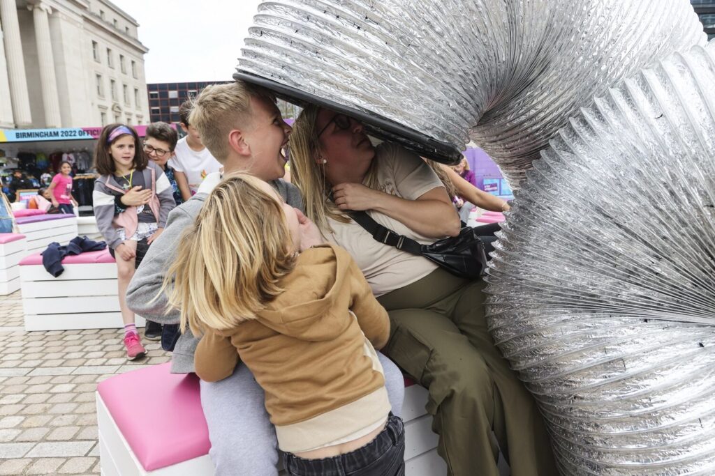 The image shows a human sized silver slinky, towering over three people as though they might go inside. The people are laughing.