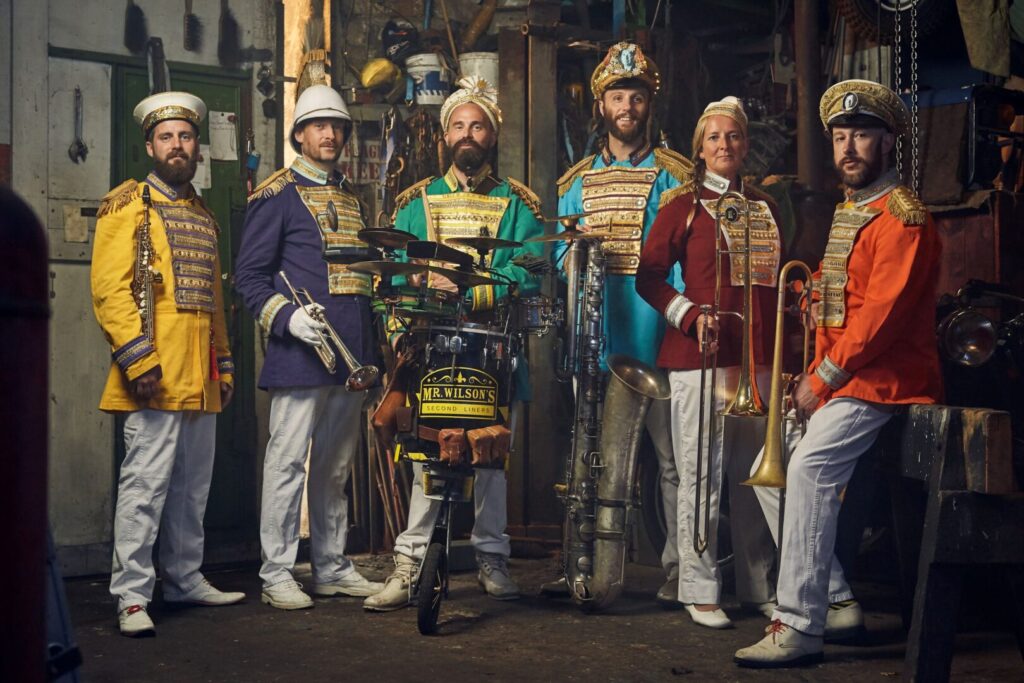 A musical group dressed in bright miltary-style clothes are facing us in a line. They have brass instruments, and one has a drum with Mr Wilson's Second Liners written on it.