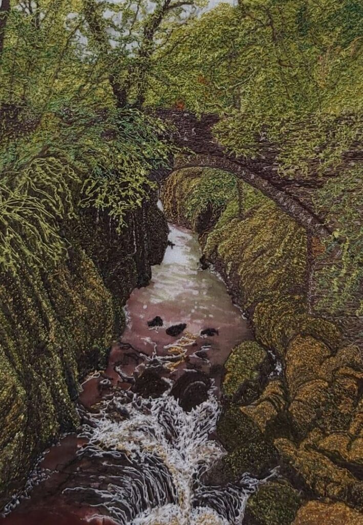 A scenic view of a narrow stream rushing through a rocky gorge, surrounded by lush greenery. An old stone bridge arches over the water, with trees and foliage framing the scene. The piece is textured by the material used to create it.