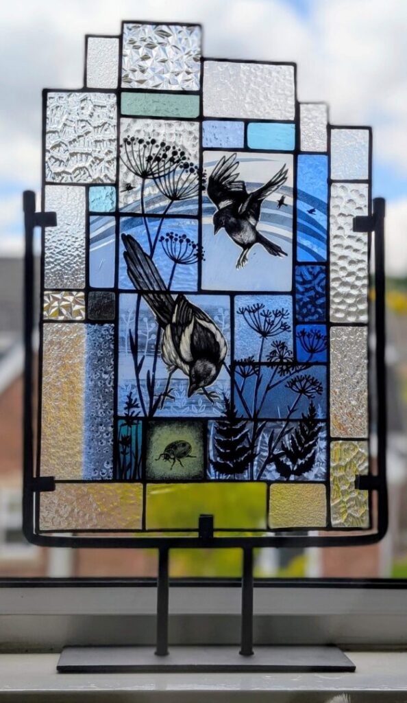 A stained glass artwork featuring two birds - one perched, one in flight - amid botanical designs. The glass is sectioned with varied textures and colours, mainly blue, clear, and yellow. Mounted on a metal stand on a windowsill, with an outdoor view behind.