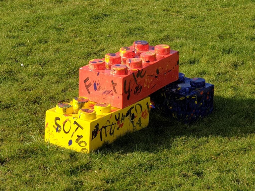 An outdoor installation, featuring three oversized toy building blocks painted in bold yellow, red, and blue. Splashed with expressive paint and graffiti-like text - e.g. “SOT 100” on the yellow block. The blocks rest on a grassy field.