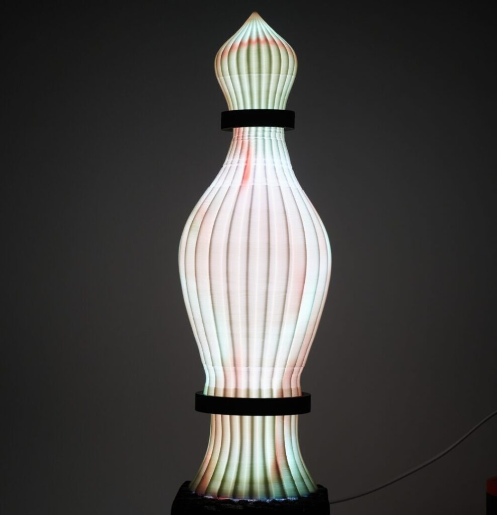 A tall, sculptural lamp with a ribbed texture and a segmented form, divided by two black bands. It glows in shades of white, green, and red, with a pointed top, rounded middle, and tapered base.