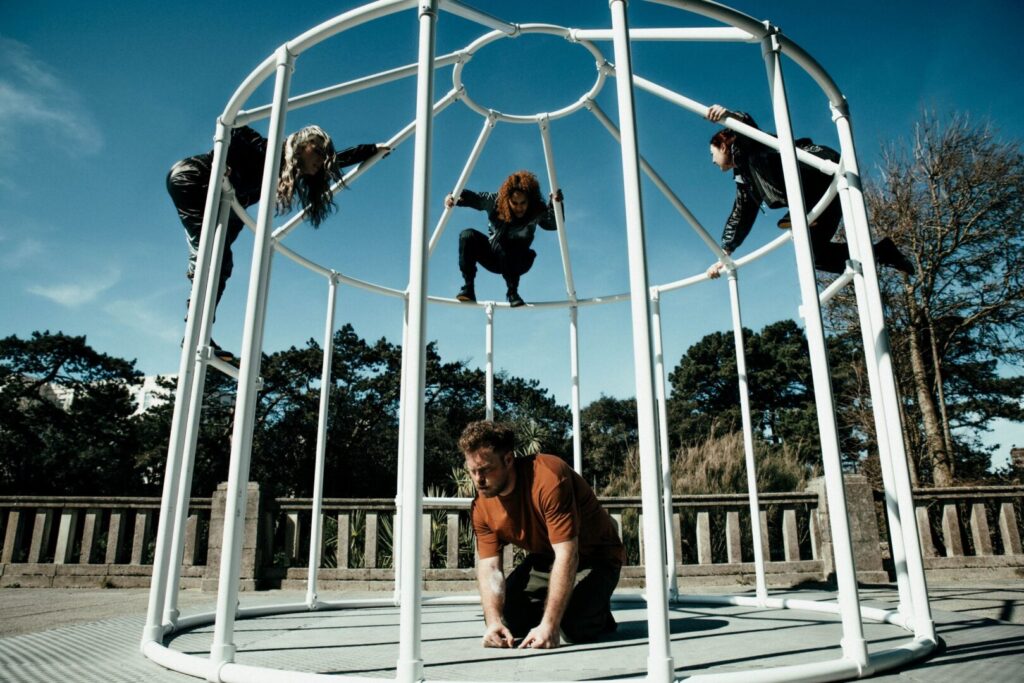 Four people are performing around a white bird cage-like structure of white bars. Three are on the outside, while one is inside on hands and knees. The cage is on a concreted are bordered by stone railings with tall trees and blue sky behind.
