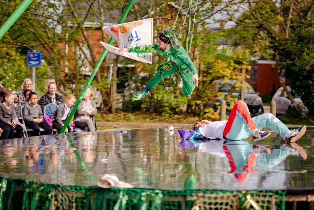 A lively outdoor performance with a man in green soaring through the air on ropes over a man in a shell suit lying on the ground. The flying performer is holding a fabric sign that is upside down and says ‘fish’ in blue above a hand-drawn picture of a goldfish. They're performing over a shiny surface, while an audience watches from the side. Trees and houses in the background show they are outdoors.