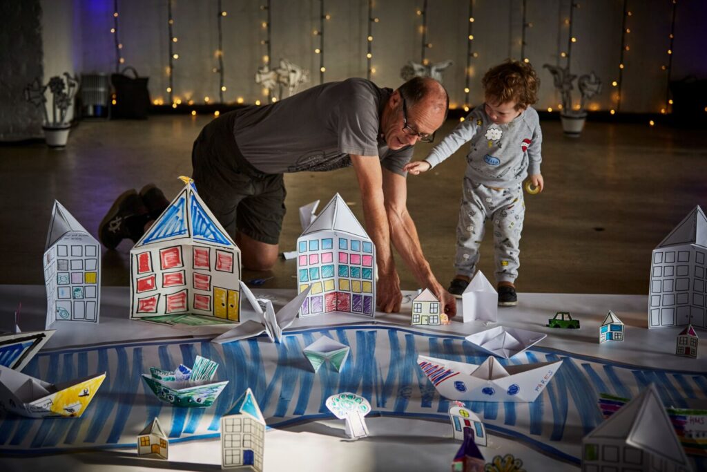 An older man on his knees and a small boy standing are behind a large paper model of a town, complete with colourful houses, boats, and trees made from folded paper. The folded city sits on a large white sheet of paper with a blue drawn river weaving through. String lights hang in the background.