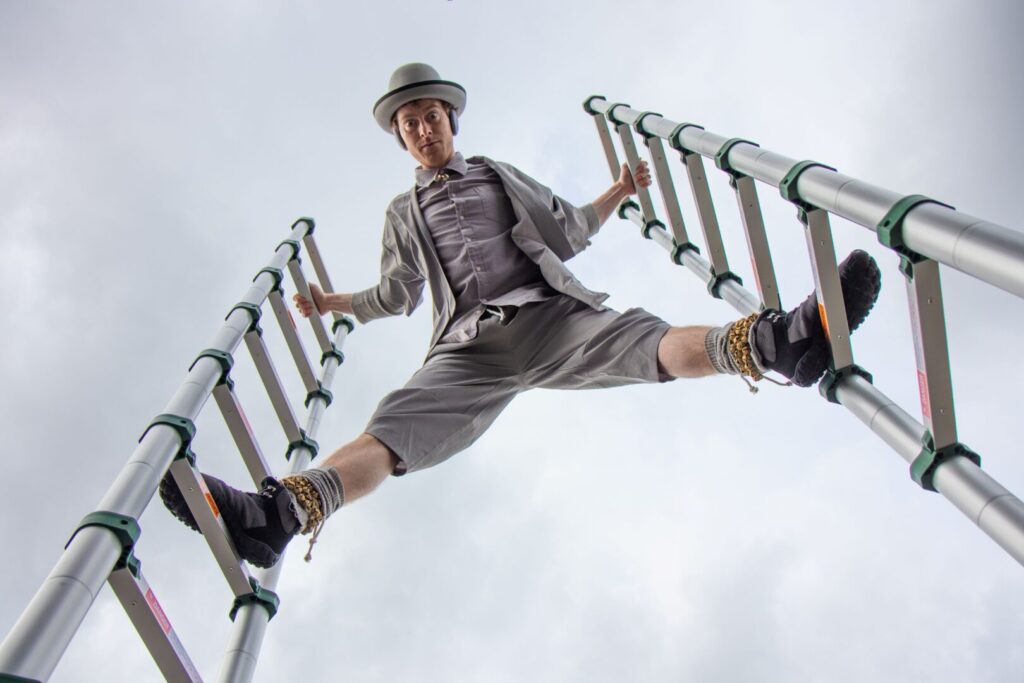 A man in a bowler hat, waistcoat and shorts pulls off a daring split between two upright ladders, one foot on each rung and hands gripping the sides. He’s looking down at us from between his wide-stretched feet, the steely grey beyond him.