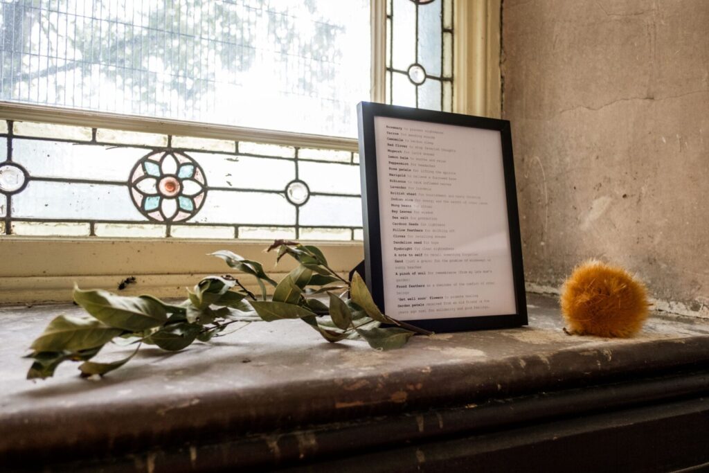 A wooden photo frame stands on a windowsill. Inside the frame s a typed list herb and their properties. The windowsill the frame sits on, the wall and window frame are clearly in an old building. The window pane has stained pattern elements to the outer borders. There is the end of a green-leafed branch and a furry, orange flower head also sitting on the windowsill.