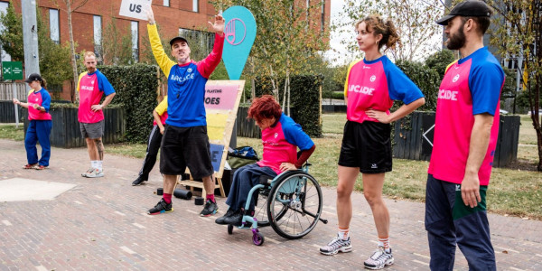 A group of performers - five standing and one in a wheelchair - are lined up in an outdoor space. They dressed in blue, pink, and yellow shirts with ARCADE written across the chest in white. One in the middle is holding up a sign that says JOIN US.