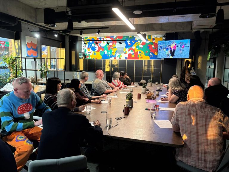 The image shows a room with a large table in the centre. Many people are sat at the table looking away from the camera and looking at a screen that has a presentation on. There are drinks and pieces of paper on the table.