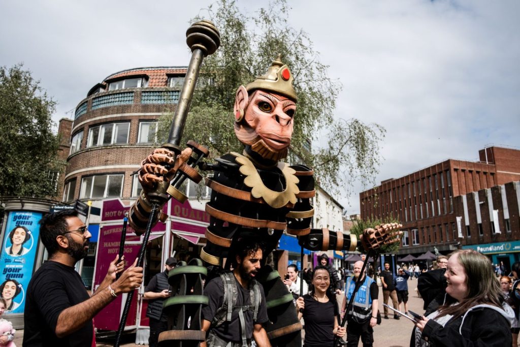 One of the characters from Ancient Giants, can be seen interacting with an audience member, who is looking at it and smiling. The character towers above them. The scene is taken on a street with buildings in the background.