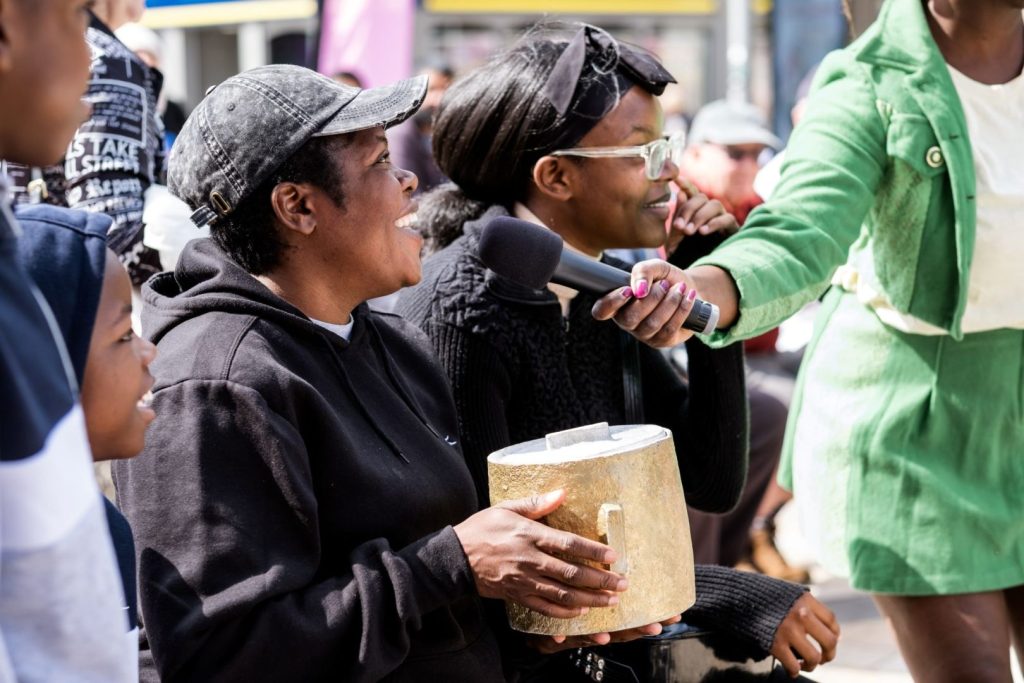 A performer in a green jacket and skirt is holding a microphone up to a member of the audience. The audience member is sitting down and is holding what looks like a large pot. the are wearing black clothing and a black cap and are smiling at the performer. Other audience members can be seen alongside.