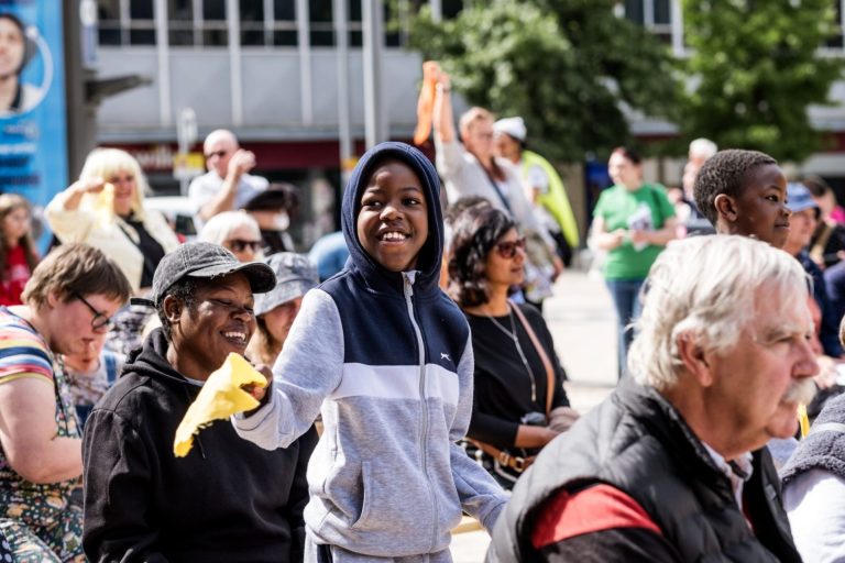 Person in a grey and navy tracksuit holding a yellow cloth during an outdoor event with people seated and standing in a sunny urban environment.